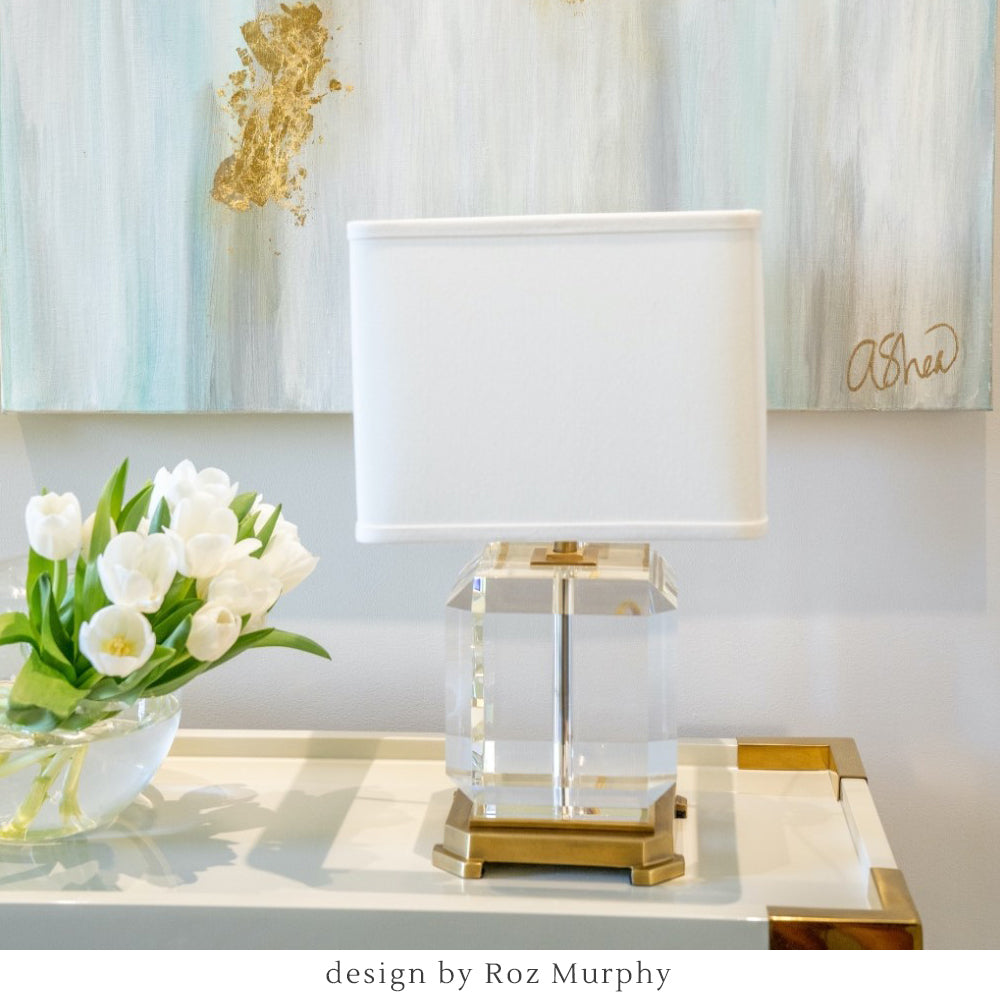 A crystal and brass table lamp with a white shade on a designer console table with flowers and a painting in the background.