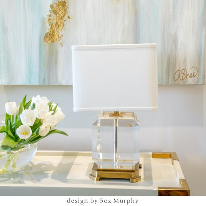 A crystal and brass table lamp with a white shade on a designer console table with flowers and a painting in the background.