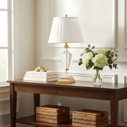 Wooden desk with a brass and crystal table lamp, books, and flowers in a bright room.