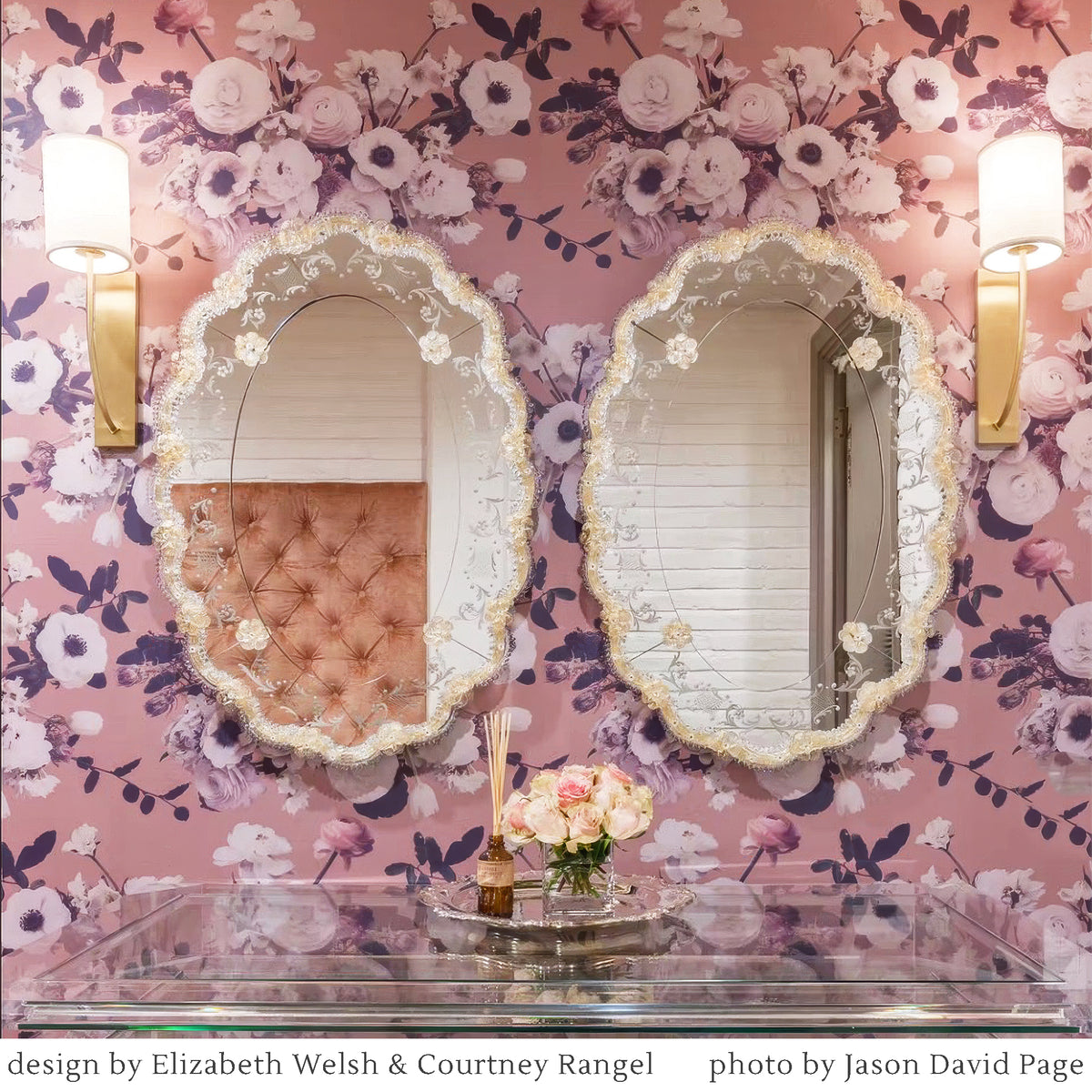 Two ornate gold Venetian mirrors with scalloped decorative trim hang on a pink floral wallpapered wall, flanked by two gold wall sconces above a glass console table with flowers.