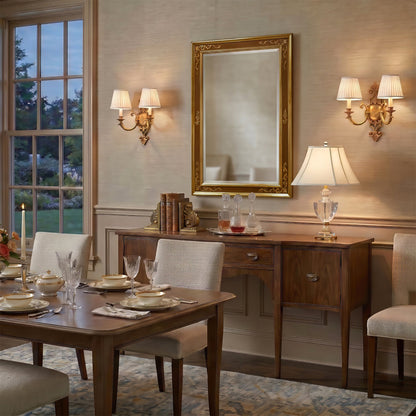 An elegant dining room scene featuring a large gold-framed rectangular mirror on a wall, flanked by two luxurious brass wall sconces. Below the mirror is a dark wood sideboard with a crystal table lamp, books, and glassware.