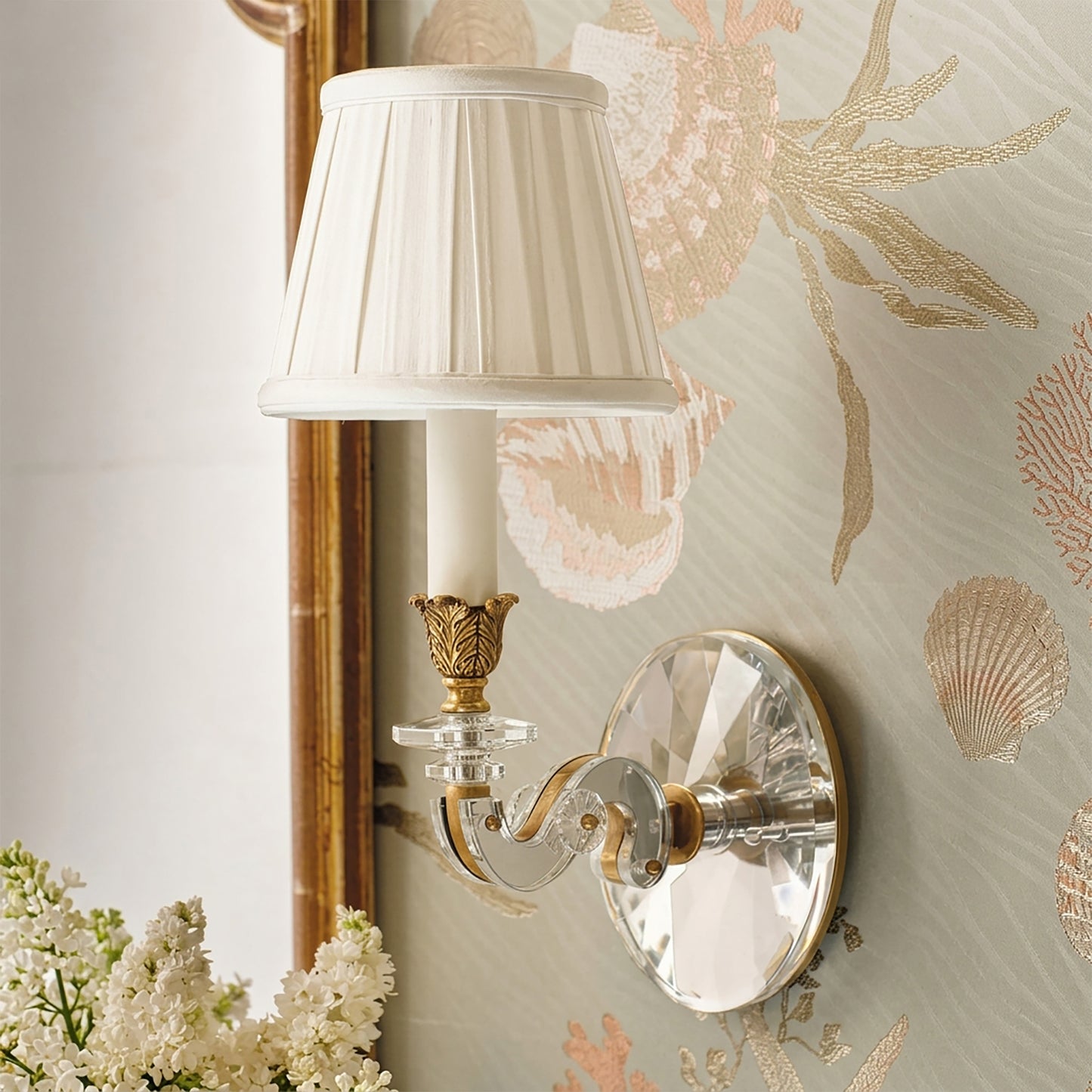 Antique brass sconce with crystal details and a white pleated shade next to a traditional mirror.