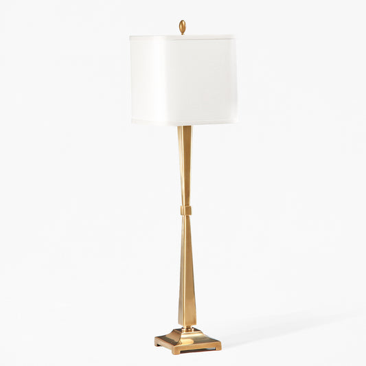 An antique brass table lamp with a square white shade against a grey background.