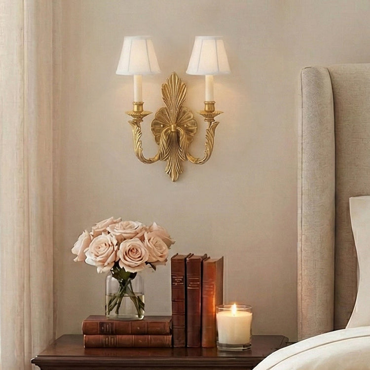 A double-light antique brass wall sconce with an ornate frame and white fabric shades, mounted on a beige bedroom wall above a nightstand with books, flowers, and a candle.