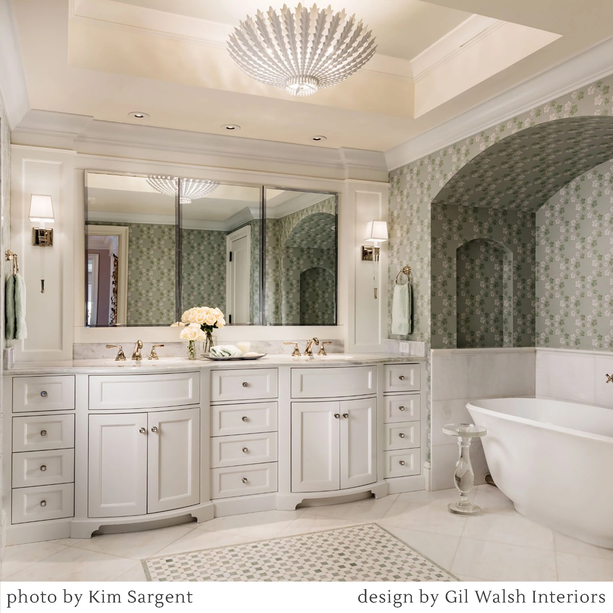 A luxury bathroom interior featuring a large white double vanity with multiple drawers and mirrors, a freestanding white bathtub, patterned wallpaper, chandelier, and two crystal sconces.