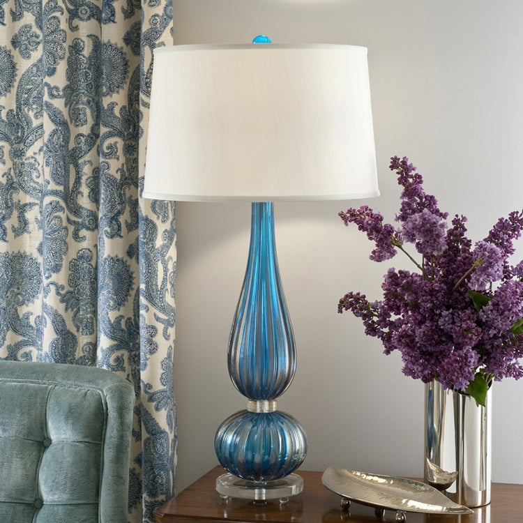 A tall blue Murano glass lamp between luxurious drapes and a large flower arrangement.