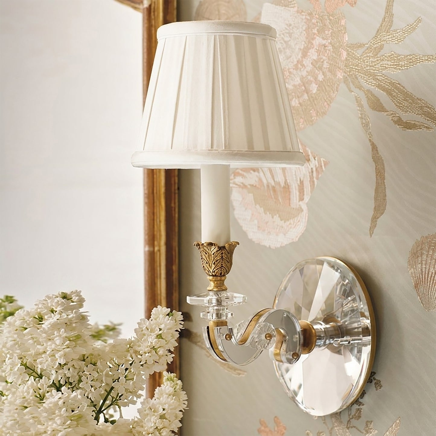 Crystal sconce with brass details and white pleated shade next to mirror.