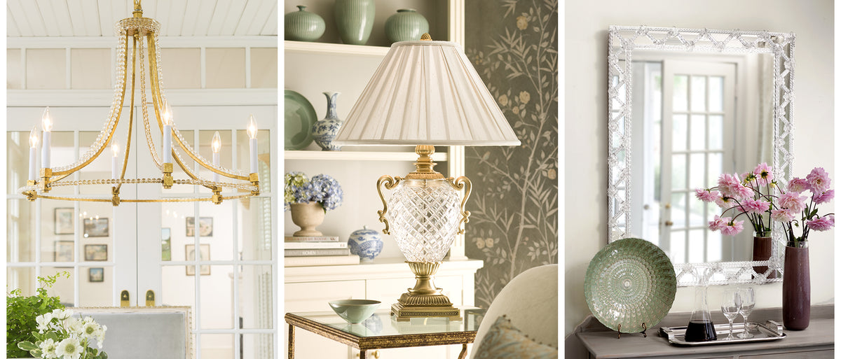 A gold chandelier in a bright room, a classic table lamp with a pleated white shade and ornate brass base, and a large Murano glass mirror above a console table with pink flowers.