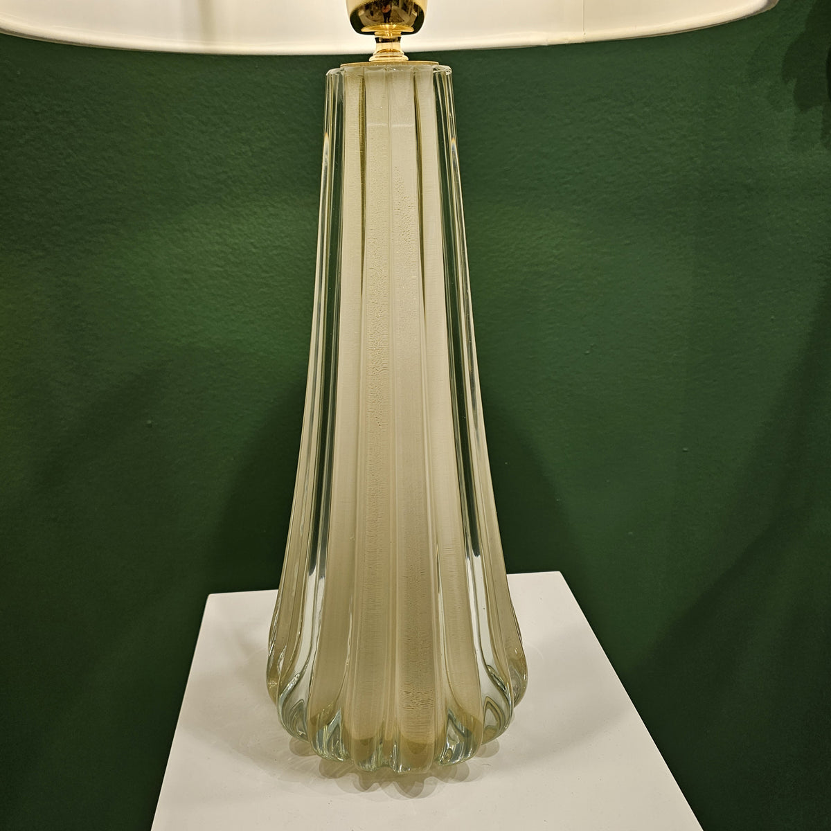 White Murano glass table lamp on a pedestal in front of a green wall.