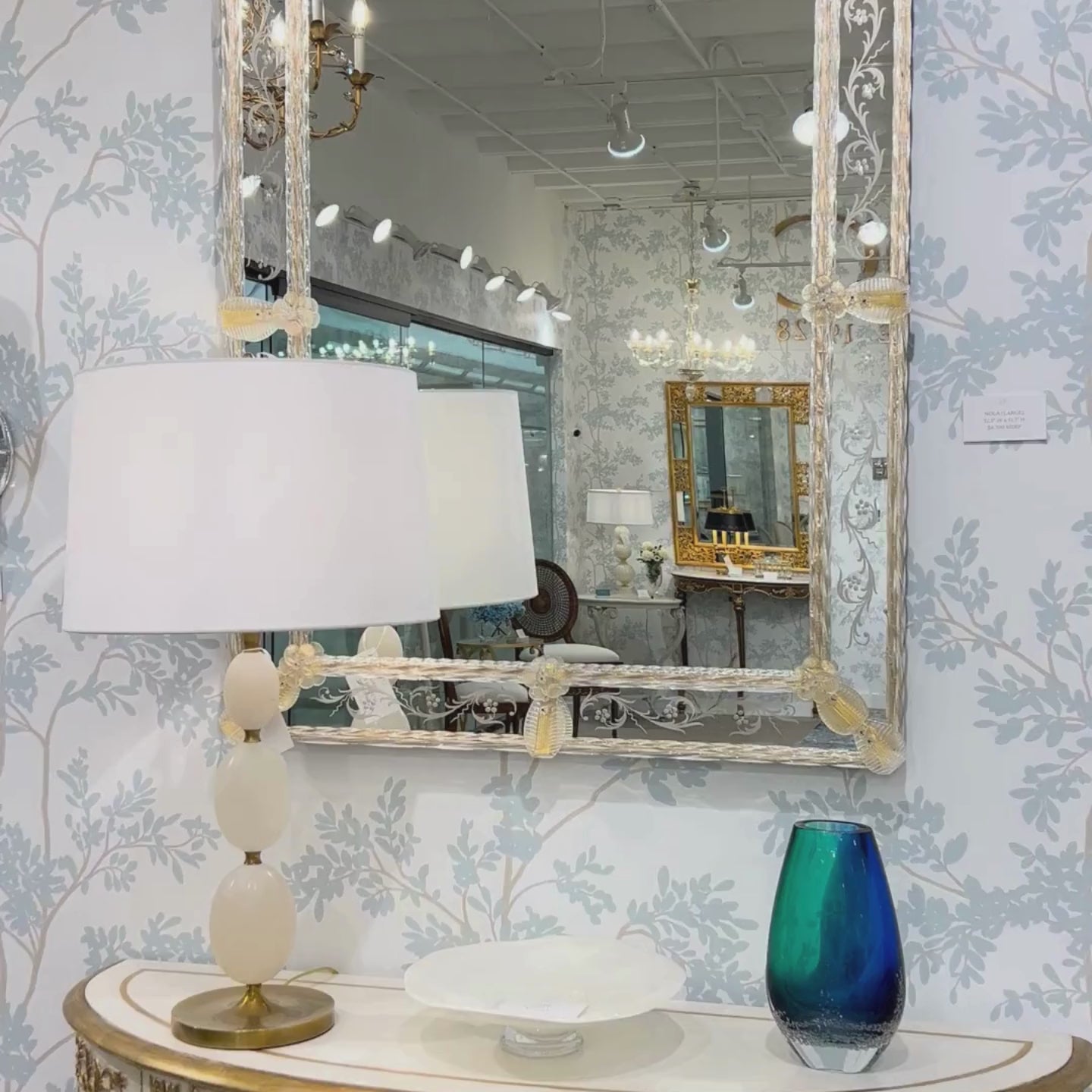 Gold Venetian glass mirror with luxurious wallpaper and brass lamp.