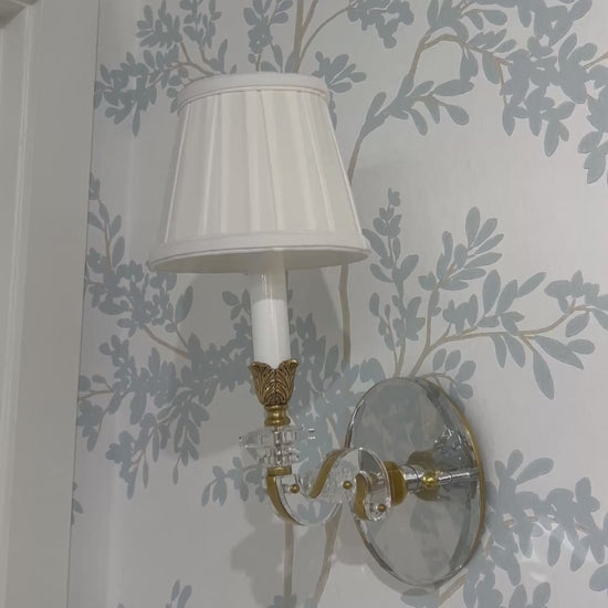 Luxurious crystal sconce with brass trim and pleated white shade on designer wallpaper.