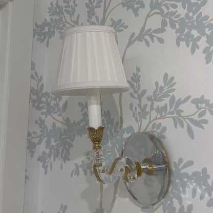 Luxurious crystal sconce with brass trim and pleated white shade on designer wallpaper.