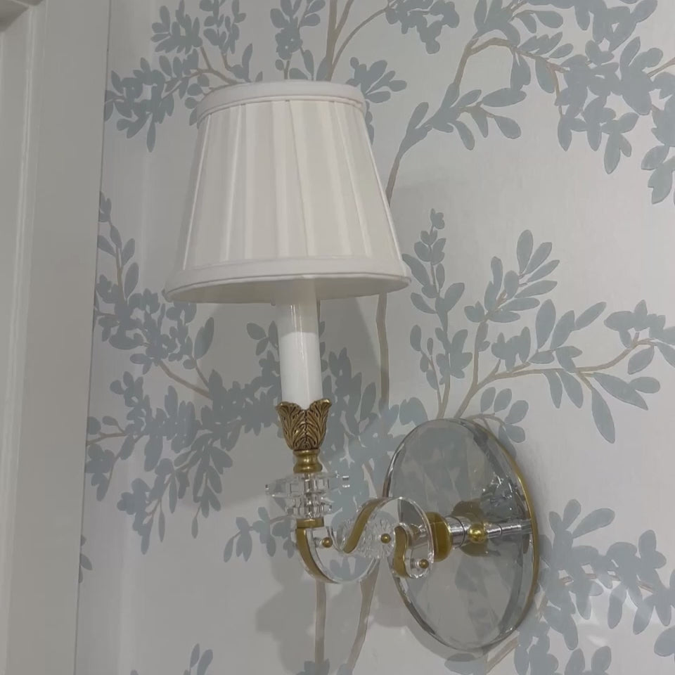 Luxurious crystal sconce with brass trim and pleated white shade on designer wallpaper.
